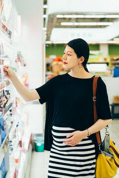 side view of young pregnant girl at shop choosing cosmetics among great variety of products. Concept of consumerism retail and purchase. future parenthood lady hold big belly and buy makeup product