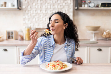 Young latin woman eating delicious pasta, enjoying tasty homemade lunch with closed eyes, sitting in kitchen