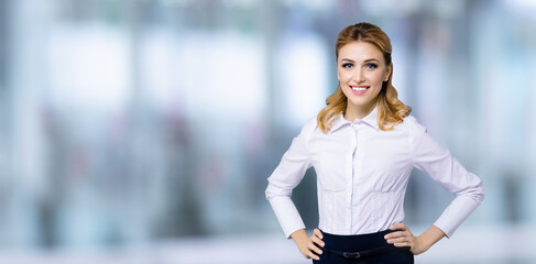 Portrait image of happy smiling businesswoman, with copy space area for text. Confident blond woman in white shirt standing in hands on hips pose, on blurred modern office background. Business concept