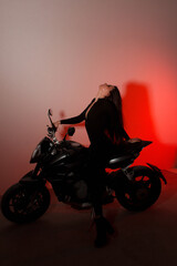 Fototapeta na wymiar teen leisure. young brunette girl with long legs sits fashion on a black sports motorcycle with closed eyes on the white wall background in red light in studio. lifestyle fashion concept, free space