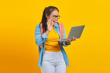 Portrait of cheerful young Asian woman student in casual wear with backpack waving to laptop during video call with friends isolated on yellow background. Education in college university concept