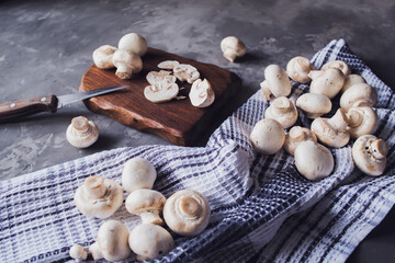 Fresh white mushrooms on a cutting board on a kitchen towel. Gray background. Knife next to champignons cut into slices. Kitchen, cooking, recipes. Mushrooms, ingredient.