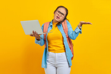 Portrait of angry young Asian woman student in casual clothes with backpack talking on smartphone while holding laptop,saying no with palms isolated on yellow background