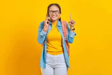 Portrait of cheerful young Asian woman student in casual clothes with backpack talking on mobile phone and pointing finger up, creating genius solutions isolated on yellow background