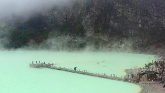 aerial drone of a green toxic sulfur lake inside a volcano called Kawah Putih with fumes and gasses rising while tourists walk a long bridge in Bandung Indonesia