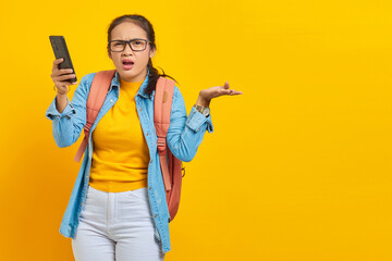 Portrait of  confused young Asian woman student in casual clothes with backpack holding mobile phone and saying no with palms isolated on yellow background.  Education in college university concept