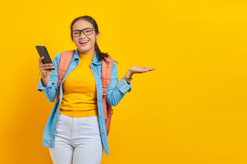 Portrait of  cheerful young Asian woman student in casual clothes with backpack using mobile phone and pointing to copy space with palms isolated on yellow background