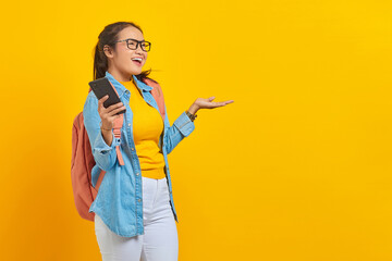 Portrait of  cheerful young Asian woman student in casual clothes with backpack using mobile phone and pointing to copy space with palms isolated on yellow background
