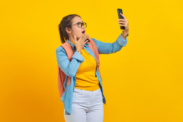 Portrait of  surprised young Asian woman student in casual clothes with backpack looking at smartphone isolated on yellow background. Education in college university concept