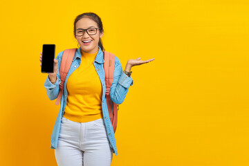 Portrait of cheerful young Asian woman student in casual clothes with backpack holding smartphone and pointing to copy space with palms isolated on yellow background