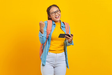 Portrait of cheerful young Asian woman student in casual clothes with backpack playing video game on mobile phone and celebrating success isolated on yellow background