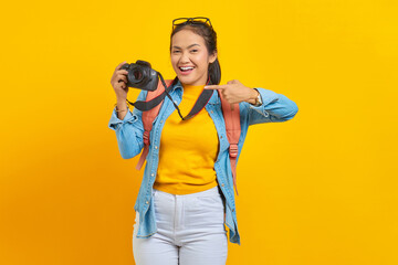 Portrait of smiling young Asian woman with backpack and finger pointing at professional camera isolated on yellow background