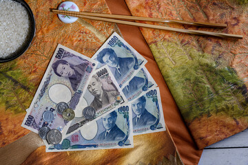 Coins and paper banknotes of Japanese yen on the background of national fabric with embroidery, a bowl of rice and chopsticks