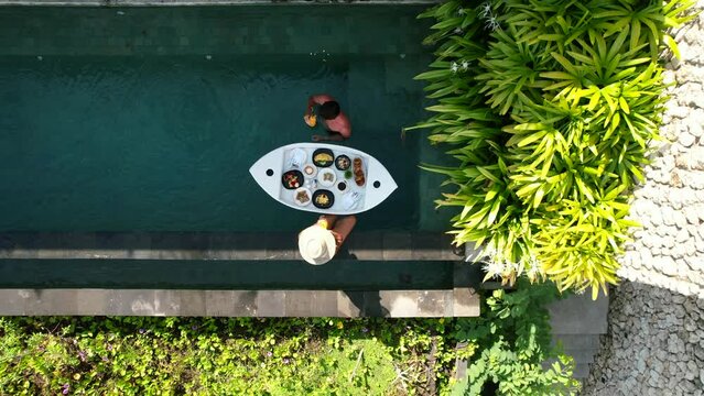 Aerial top down view of young couple with a floating breakfast poolside at a tropical luxury villa during vacation in Bali Indonesia