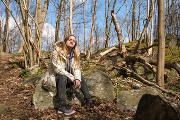 Naklejka premium Young woman is sitting on huge stone in spring park. Sunny day. Naked trees. Rest.