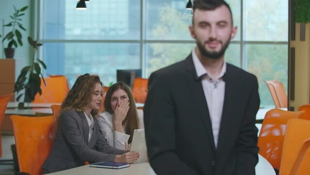 Beautiful Caucasian women gossiping sitting at table in office discussing blurred man at front. Cheerful colleagues sharing rumors looking at coworker indoors. Employment and fun concept