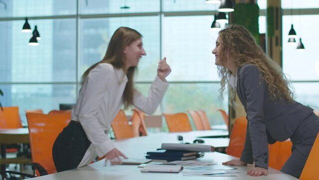 Young women whispering on ear gossiping in office indoors laughing. Side view portrait of beautiful carefree Caucasian female colleagues sharing rumors at workplace indoors smiling leaning on table