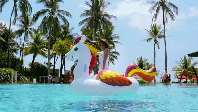 Happy travel vacation woman on inflatable unicorn float mattress dancing and having fun in swimming pool in luxury tropical hotel. Female tourist in swimwear relaxing sunbathing in resort