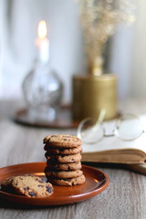 Stack of chocolate chip cookies, open book and reading glasses, lit candle and vase with flowers on the table. Hygge at home. Selective focus.