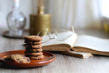 Fototapeta na wymiar Stack of chocolate chip cookies, open book and reading glasses, lit candle and vase with flowers on the table. Hygge at home. Selective focus.