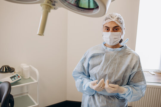 Portrait Of Female Doctor Surgeon In Blue Scrubs Putting On Surgical Gloves And Looking At Camera