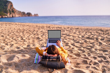 Millennial freelancer female using mockup laptop with white screen blank and lying on the sand...