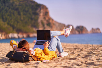 Millennial freelancer female using mockup laptop with empty screen blank and lying on the sand...