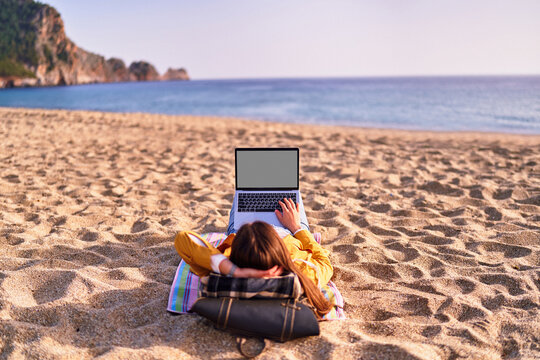 Millennial freelancer female using mockup laptop with white screen blank and lying on the sand beach by the sea. Dream office remote work concept