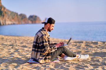 Young happy joyful carefree satisfied millennial freelancer man using laptop on sand beach by the sea. Dream office work concept