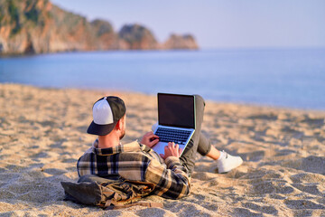 Millennial freelancer guy using mockup laptop with empty screen blank and lying on the sand beach by the sea. Dream office remote work concept - obrazy, fototapety, plakaty