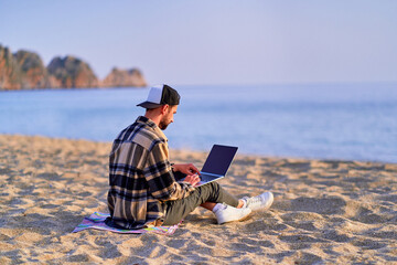 Young happy joyful carefree satisfied millennial freelancer man using laptop on sand beach by the...