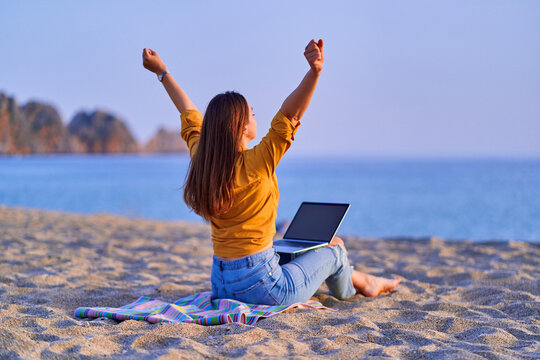 Satisfied millennial free freelancer woman with open arms using computer and sitting on sand beach by the sea. Enjoyment of dream office remote work concept