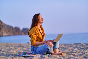 Satisfied millennial free freelancer woman using computer and sitting on sand beach by the sea....