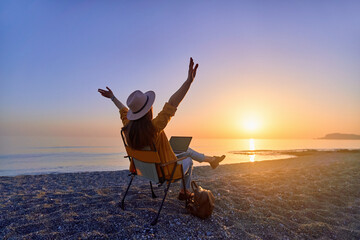 Satisfied free millennial freelancer woman with open arms using computer and sitting on the beach...