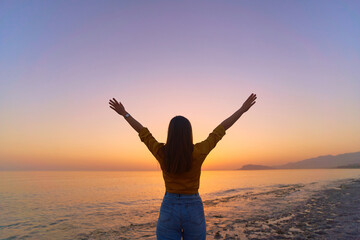 Fototapeta premium Back view of free calm bliss satisfied woman standing with open arms against of sunset sky by the sea in a happy beautiful inspired moment of her life