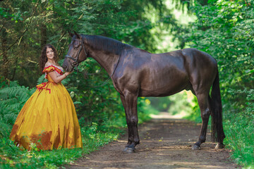 A young woman in a vintage yellow dress walks with a brown horse in a green park on a summer day