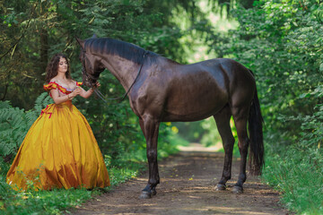 A young woman in a vintage yellow dress walks with a brown horse in a green park on a summer day