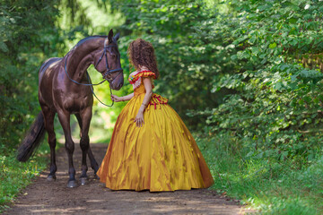 A young woman in a vintage yellow dress walks with a brown horse in a green park on a summer day