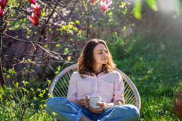 Young beautiful woman enjoying spring time while sitting in blooming garden of her home with a cup...