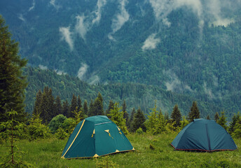 Summer view of small comfortable modern tourist tent on grassy hill under beautiful blue sky on foggy mountains covered with forest background.