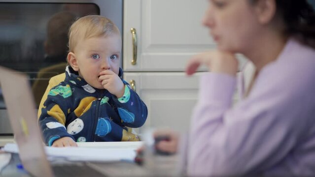 Working woman with one year old baby boy in high chair, mother can free her hands to go about her business. Mother with child works on laptop at home. High quality 4k footage