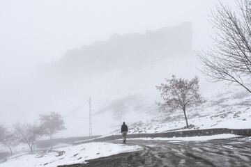 Huge rock  hill behind foggy air in winter and snow, man is walking on the path, there are trees