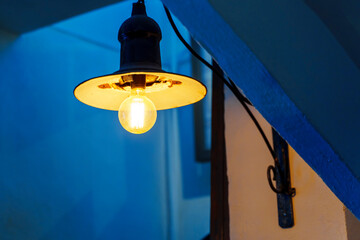 An antique lamp under a tin lampshade shines with a yellow light on the blue walls
