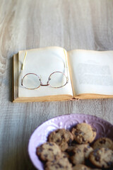 Fototapeta premium Bowl of chocolate chip cookies, open book and reading glasses on the table. Selective focus.