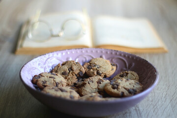 Bowl of chocolate chip cookies, open book and reading glasses on the table. Selective focus.