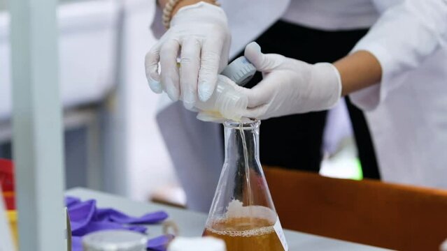Scientist pours brown reagent from plastic bottle into glass flask in laboratory. Worker in rubber gloves does scientific research close view