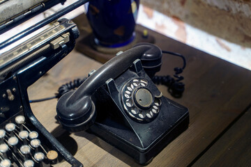 Old dust-covered black phone with disk dialing on a wooden table next to the typewriter