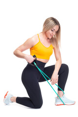 A beautiful, athletic, slim, smiling and cheerful woman in an orange top and black sweatpants performs exercises on the back muscles with a green rubber band while sitting on one knee. Lifestyle