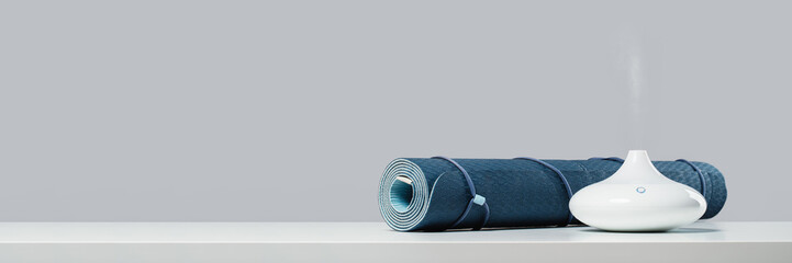 Rolled blue yoga mat and essential oil diffusor for enhanced practice. Ultrasonic and humidifying aromatherapy. Meditation fitness and exercise banner copy space. Active lifestyle. home or gym club - obrazy, fototapety, plakaty