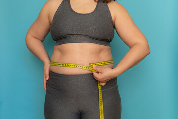 Unrecognizable fat plump plus-size overweight woman standing in grey sports bra, leggings, showing excess naked belly, measuring waist with tape on blue background. Dieting, unhealthy food, obesity.